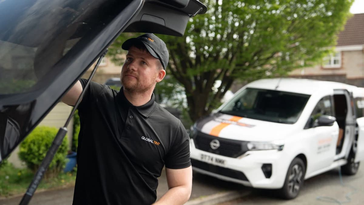 Global Tint installer working beneath an open car hatch with a branded service van behind him